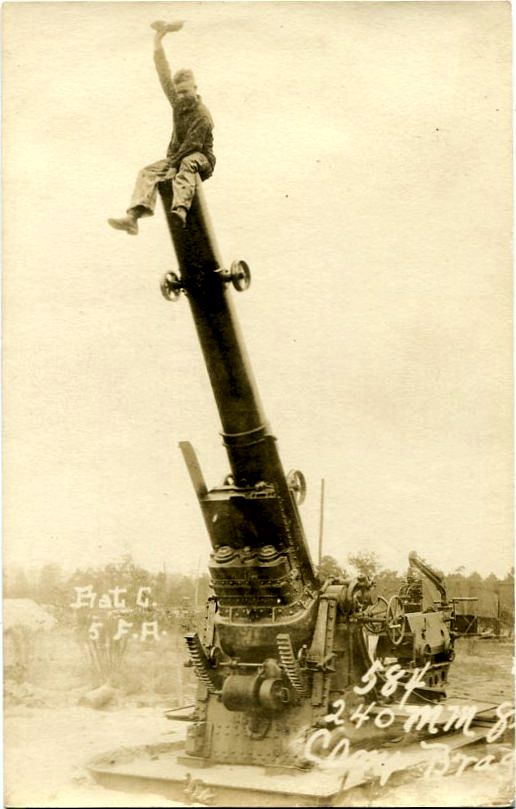 Real-photo postcard of a photograph of an unidentified U.S. Army soldier with Battery C, 5th Field Artillery Regiment, sitting on the end of the raised-up barrel of a M1918 240mm howitzer gun at Camp Bragg, N.C., taken between 1918 and 1922 [circa 1918-1922].