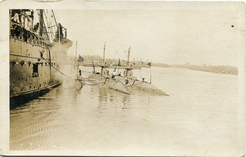 Real-photo postcard of an unidentified troop transport ship [believed to be the USS Powhatan, a U.S. Navy troop transport ship] parked in an unidentified harbor [believed to be in France], with three U.S. Navy submarines alongside the ship tied to the ship with ropes. Two of the three submarines have American flags flying from their bows. Image taken during World War I (Undated).